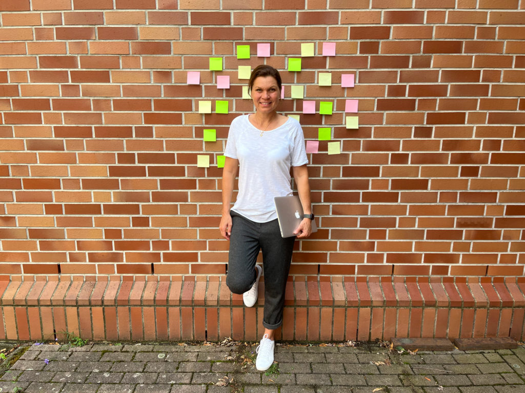 Louise Hansen stehend vor einer Hauswand, an der verschiedenfarbige Post-its kleben. In ihrer Hand hält sie einen Laptop.